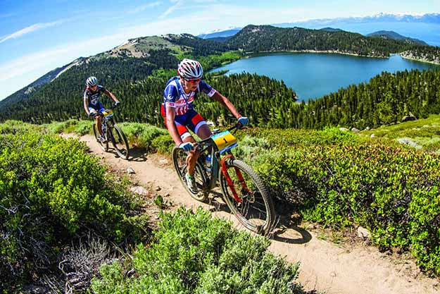 Two people biking across a mountain with a forest in the background looking out onto a lake.